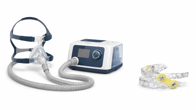 A CPAP machine with mask and hose shown beside a small custom oral appliance at the same scale