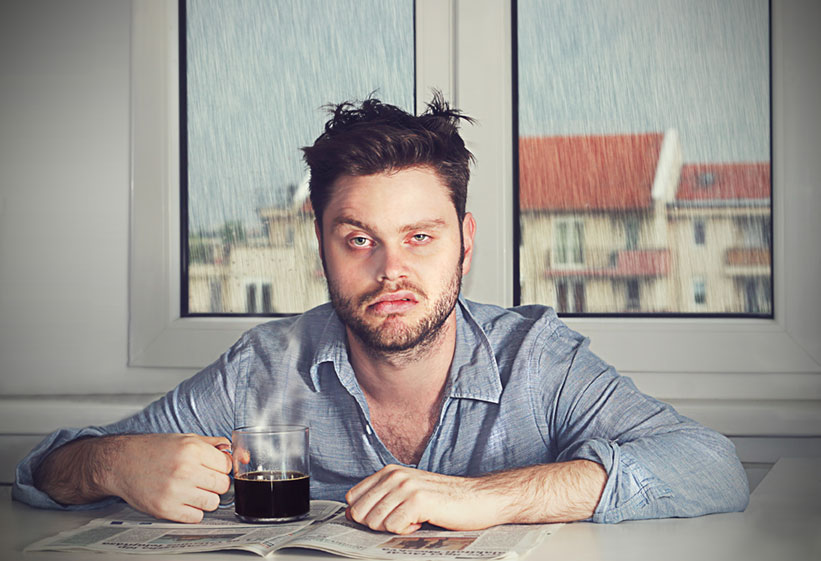 Tired man sitting at a table with a scowl on his face, newspaper open, and drinking coffee